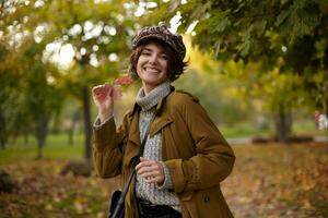 Extérieur coup de content Jeune attrayant brunette femelle habillé dans élégant porter souriant largement à caméra tandis que posant plus de parc Contexte avec feuille dans élevé main photo