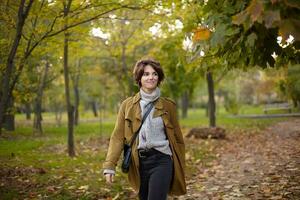 Extérieur coup de positif attrayant Jeune marron aux cheveux femelle portant élégant confortable vêtements tandis que en marchant plus de jauni des arbres dans parc, étant dans agréable ambiance et souriant agréablement photo