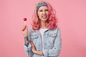 Jeune agressif magnifique rose aux cheveux femme dans denim monsieur, en portant une sucette, regards à le caméra et dénudé le sien dents, des stands plus de rose Contexte. photo