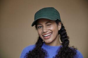 fermer de de bonne humeur Jeune frisé brunette femme avec foncé peau un clin d'oeil Heureusement à caméra, en gardant sa longue cheveux dans tresses, permanent plus de beige Contexte dans violet chandail et vert base-ball casquette photo