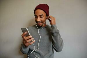 horizontal coup de attrayant Jeune barbu Masculin avec foncé peau en train de regarder vidéos avec téléphone intelligent plus de blanc mur, habillé dans gris chandail et Bourgogne casquette photo