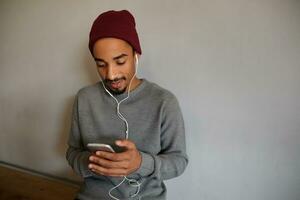 intérieur coup de jolie concentré Jeune foncé écorché homme avec barbe en portant téléphone intelligent et vérification boites aux lettres tandis que séance plus de blanc mur dans décontractée vêtements photo