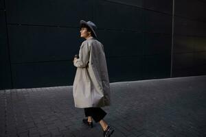 à la mode Jeune jolie frisé femme avec court la Coupe de cheveux en marchant sur ville rue dans beige longue tranchée, noir pantalon et large gris chapeau, porter à emporter café pour sa collègue photo