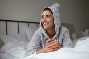 portrait de de bonne humeur attrayant Jeune foncé aux cheveux femme dans gris confortable vêtements mensonge sur lit et à la recherche positivement de côté, pliant mains et souriant Heureusement photo