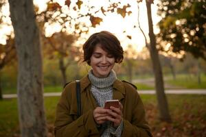 positif Jeune attrayant marron aux cheveux femme avec Naturel maquillage en portant téléphone intelligent dans élevé mains et souriant gaiement tandis que vérification sa messages, posant plus de jauni des arbres photo