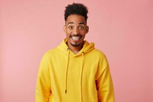 Jeune content étonné africain américain gars dans Jaune sweatshirt à capuche, entendu le nouvelles cette le sien préféré bande est à venir à le sien ville avec une concert, largement souriant et à la recherche à le caméra plus de rose Contexte. photo