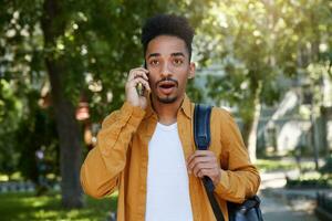 Jeune africain américain homme en marchant dans le parc, porte dans une Jaune chemise et une blanc T-shirt, parlant sur le téléphone, entend incroyable nouvelles, regards surpris à le gadget. photo