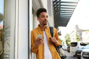 portrait de Jeune calme foncé écorché homme dans Jaune chemise en marchant vers le bas le rue , détient Téléphone, bavardage avec petite amie, regards pensivement. photo