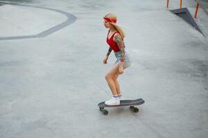 Jeune magnifique tatoué Dame avec blond cheveux dans une rouge T-shirt et denim shorts, avec une tricoté bandana sur sa diriger, profiter le journée et raids sur planche à roulette dans patin parc. photo