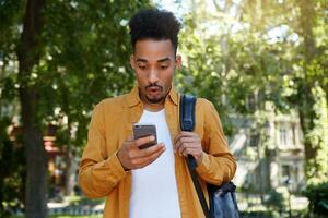 Jeune africain américain gars en marchant dans le parc, porte dans une Jaune chemise et une blanc T-shirt, eu une message de une ami avec incroyable nouvelles, regards surpris à le gadget. photo