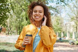 Jeune mignonne frisé foncé écorché fille largement souriant, en portant une tasse de café, portant une Jaune veste, marche dans le parc, écoute à musique, et profiter le temps. photo