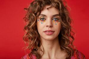 studio coup de attrayant Jeune frisé femme avec multicolore points sur sa visage à la recherche à caméra avec lumière sourire, permanent plus de rouge Contexte avec calme visage photo