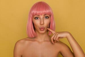 fermer de Jeune jolie aux yeux bleus rose aux cheveux femme avec branché bob la Coupe de cheveux faire la moue sa lèvres tandis que à la recherche à caméra et en gardant indice doigt sur sa joue, isolé plus de moutarde Contexte photo