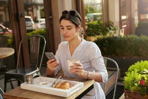 attrayant Jeune foncé aux cheveux femme avec des lunettes de soleil sur sa tête en portant mobile téléphone dans sa main et à la recherche sur écran avec plié lèvres tandis que ayant tasse de café photo