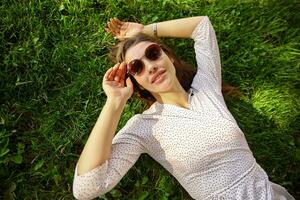 Extérieur portrait de magnifique Jeune longue aux cheveux brunette Dame portant des lunettes de soleil et blanc à pois robe tandis que relaxant plus de vert herbe avec élevé mains, à la recherche calmement à caméra photo