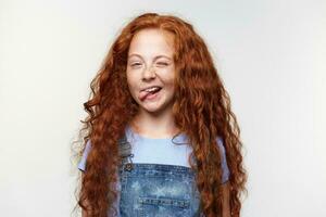 portrait de marrant mignonne taches de rousseur peu fille avec gingembre cheveux, clins d'oeil et spectacles langue à le caméra, des stands plus de blanc Contexte. photo