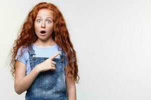 portrait de demandé mignonne taches de rousseur peu fille avec gingembre cheveux, veut à dessiner vous attention à le copie espace sur le droite côté et points avec des doigts, des stands plus de blanc mur avec large ouvert bouche. photo