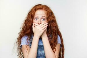 portrait de effrayé mignonne taches de rousseur peu fille avec gingembre cheveux, entend entendu une terrible histoire, couvert bouche avec palmiers, des stands plus de blanc mur avec large ouvert yeux avec peur expression. photo