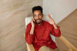 intérieur coup de Jeune Beau court aux cheveux barbu foncé écorché gars séance dans chaise tandis que ayant téléphone conversation et élevage émotionnellement le sien main, posant plus de Accueil intérieur photo