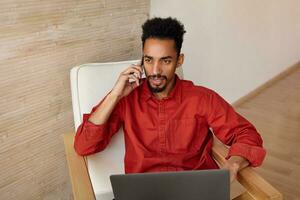 perplexe Jeune aux yeux bruns foncé écorché homme avec court la Coupe de cheveux en gardant mobile téléphone dans élevé main tandis que fabrication appel, travail dans confortable chaise plus de beige intérieur photo