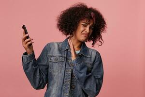 portrait de mécontent Jeune frisé brunette femelle avec foncé peau en mouvement une façon de se combiné et à la recherche à caméra avec moue, habillé dans décontractée porter tandis que permanent plus de rose Contexte photo