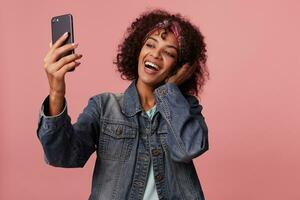 joyeux charmant souriant foncé écorché frisé brunette Dame avec court la Coupe de cheveux portant coloré Bandeau tandis que posant plus de rose arrière-plan, élevage main avec mobile téléphone tandis que fabrication photo de se