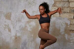 studio photo de Jeune svelte foncé écorché frisé brunette femme dans noir Haut et beige leggins en train de préparer moderne Danse tandis que posant plus de brique mur Contexte