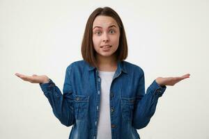 perplexe Jeune charmant aux yeux bleus brunette femme avec bob la Coupe de cheveux élevage confusément mains tandis que à la recherche avec surprise à caméra, permanent plus de blanc Contexte photo