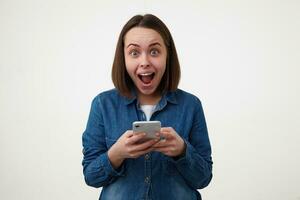 excité Jeune charmant foncé aux cheveux femme avec décontractée coiffure à la recherche hébété à caméra avec large bouche ouvert tandis que permanent plus de blanc Contexte avec sa téléphone intelligent photo