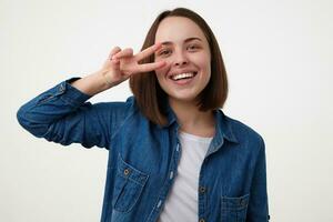 de bonne humeur Jeune attrayant foncé aux cheveux femme souriant volontier à caméra et en gardant paix geste près sa affronter, permanent plus de blanc Contexte dans décontractée vêtements photo