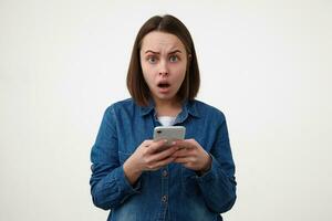 intérieur coup de sous le choc Jeune foncé aux cheveux Dame avec Naturel maquillage à la recherche étonné à caméra tandis que en gardant sa téléphone intelligent dans élevé mains, posant plus de blanc Contexte photo