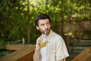 Extérieur coup de Jeune jolie Masculin avec barbe séance plus de vert des arbres sur banc, à la recherche de côté et en buvant limonade, portant décontractée vêtements photo