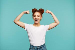 gaiement Jeune attrayant roux femelle dans décontractée vêtements élevage mains tandis que démontrant sa biceps et souriant Heureusement tandis que permanent plus de bleu Contexte photo