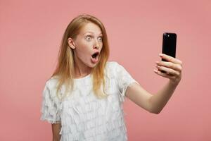 sous le choc Jeune attrayant femme avec rusé cheveux élevage main avec téléphone intelligent dans il et à la recherche à caméra avec large yeux et bouche ouvert, isolé plus de rose Contexte photo