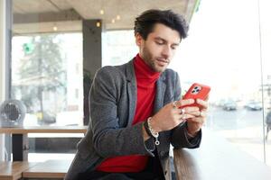 élégant Jeune jolie foncé aux cheveux barbu homme en gardant mobile téléphone dans le sien mains et à la recherche avec attention sur écran tandis que étant isolé plus de Urbain Contexte photo