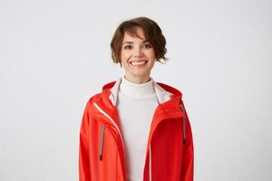 portrait de Jeune largement souriant mignonne court aux cheveux fille habillé dans blanc le golf et rouge pluie manteau, à la recherche à le caméra avec une content expression, permanent plus de blanc Contexte. photo