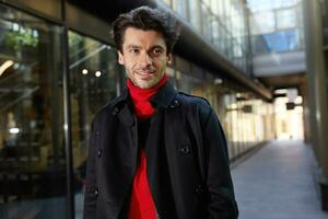 portrait de Jeune jolie marron aux cheveux barbu gars souriant gaiement tandis que en marchant Extérieur sur ensoleillé jour, habillé dans formel vêtements tandis que posant plus de ville Contexte photo