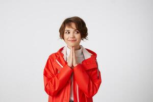 agréable Jeune court aux cheveux Dame dans blanc le golf et rouge pluie manteau, avec en coupe mains à la recherche loin, souriant et en pensant à propos quelque chose savoureux. permanent plus de blanc Contexte. photo