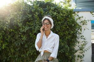 magnifique brunette femme dans branché vêtements et ancien lunettes posant plus de vert des buissons sur chaud ensoleillé jour, ayant agréable conversation sur téléphone et souriant gaiement photo