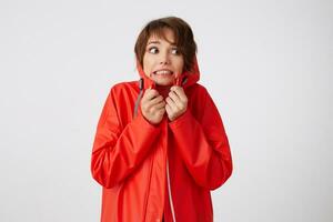 portrait de mignonne Jeune effrayé court aux cheveux femme dans rouge pluie manteau, renfrogné et craignant à la recherche à le droite, se sent froid, cache dans le capot. permanent plus de blanc Contexte. photo