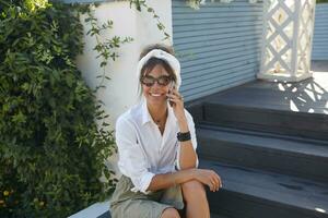 Extérieur photo de magnifique de bonne humeur brunette femme dans branché vêtements et rétro lunettes séance plus de été terrasse sur chaud ensoleillé jour, ayant agréable parler sur téléphone et souriant joyeusement