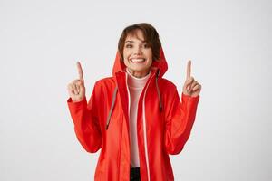 mignonne positif court aux cheveux fille dans rouge pluie manteau, largement sourires, regards à le caméra, veut à dessiner votre attention à le copie espace au dessus sa diriger, points les doigts en haut. permanent plus de blanc Contexte. photo