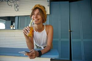 de bonne humeur Jeune brunette Dame avec décontractée coiffure portant blanc Haut et moutarde Bandeau et en buvant limonade avec paille, à la recherche devant rêveusement tandis que permanent plus de café intérieur photo