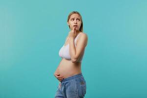 effrayé Jeune blond longue aux cheveux femme étant Enceinte tandis que permanent plus de bleu arrière-plan, renfrogné les sourcils tandis que à la recherche avec peur de côté et étreindre sa ventre photo