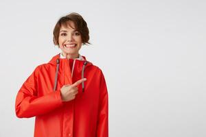 Jeune largement souriant mignonne court aux cheveux Dame habillé dans blanc le golf et rouge pluie manteau, veut à dessiner votre attention à le copie espace, points les doigts à le droite. permanent plus de blanc Contexte. photo