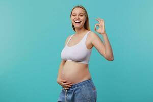 de bonne humeur Jeune jolie à tête blanche femelle montrant bien terminé signe et donnant Heureusement clin d'œil à caméra, habillé dans blanc sportif Haut et jeans tandis que posant plus de bleu Contexte photo