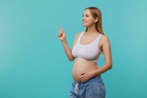 côté vue de Jeune charmant blond longue aux cheveux femelle avec Naturel maquillage en gardant sa index élevé tandis que à la recherche gaiement devant et émouvant sa ventre, Aller à être maman bientôt photo