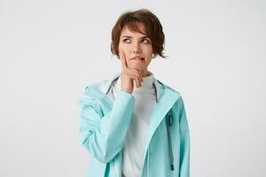 portrait de Jeune pensif cheveux courts femme dans blanc le golf et lumière bleu pluie manteau, touche joue et mord lèvres, regards loin, des stands plus de blanc Contexte. photo