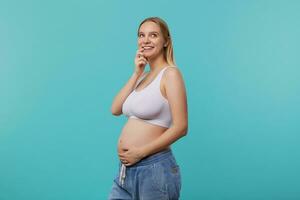 intérieur photo de Jeune Enceinte blond longue aux cheveux femme en portant sa ventre tandis que à la recherche pensivement vers le haut avec agréable sourire, permanent plus de bleu Contexte