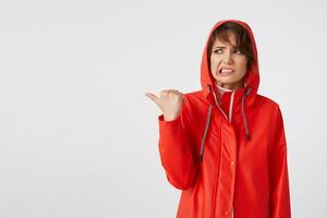 Jeune malheureux mignonne court aux cheveux Dame habillé dans rouge pluie manteau, à la recherche avec dégoûter, veut à dessiner votre attention à le copie espace, points les doigts à le gauche. permanent plus de blanc Contexte. photo